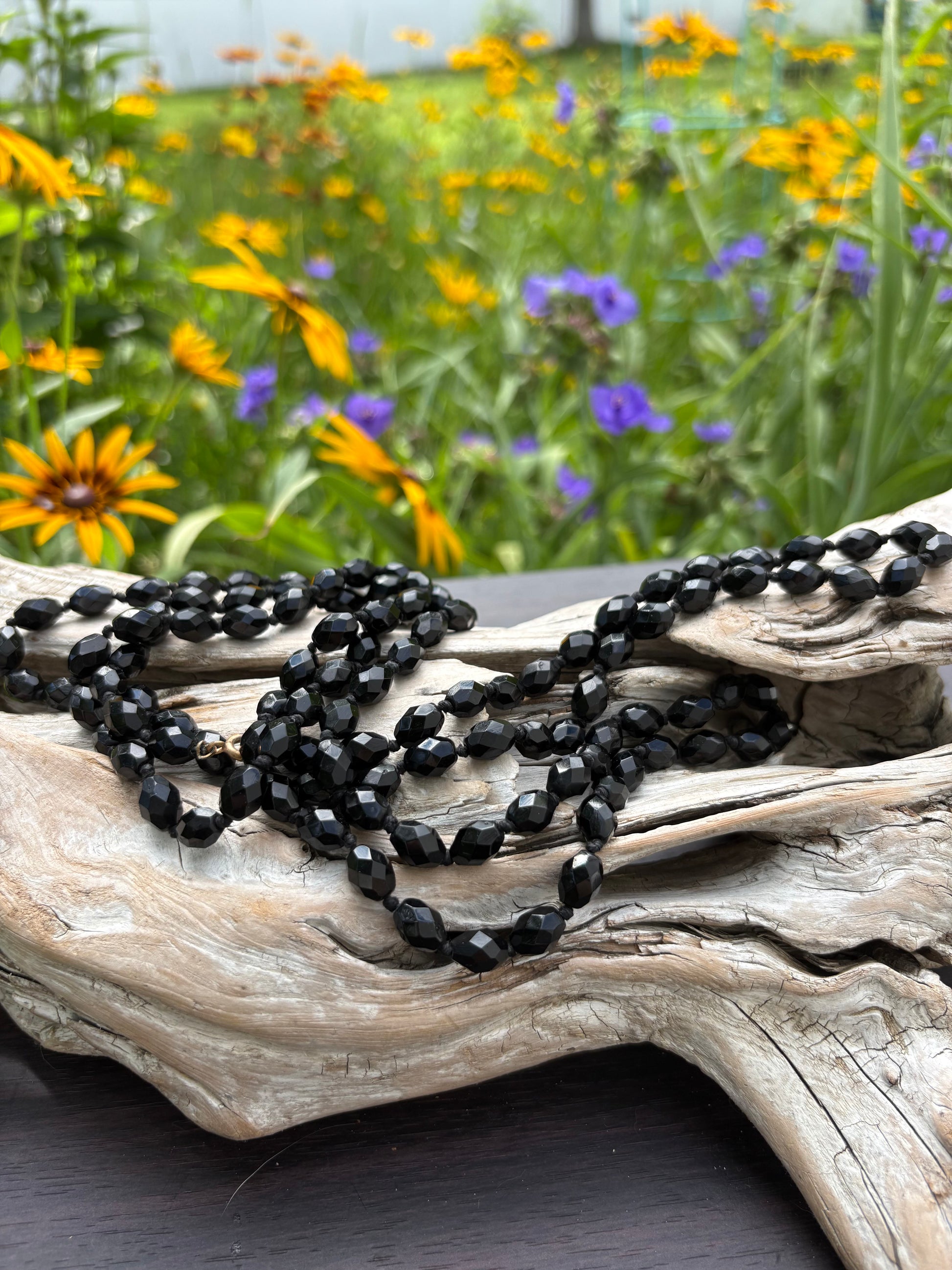 Black beaded necklace draped over a textured wooden surface with flowers in the background