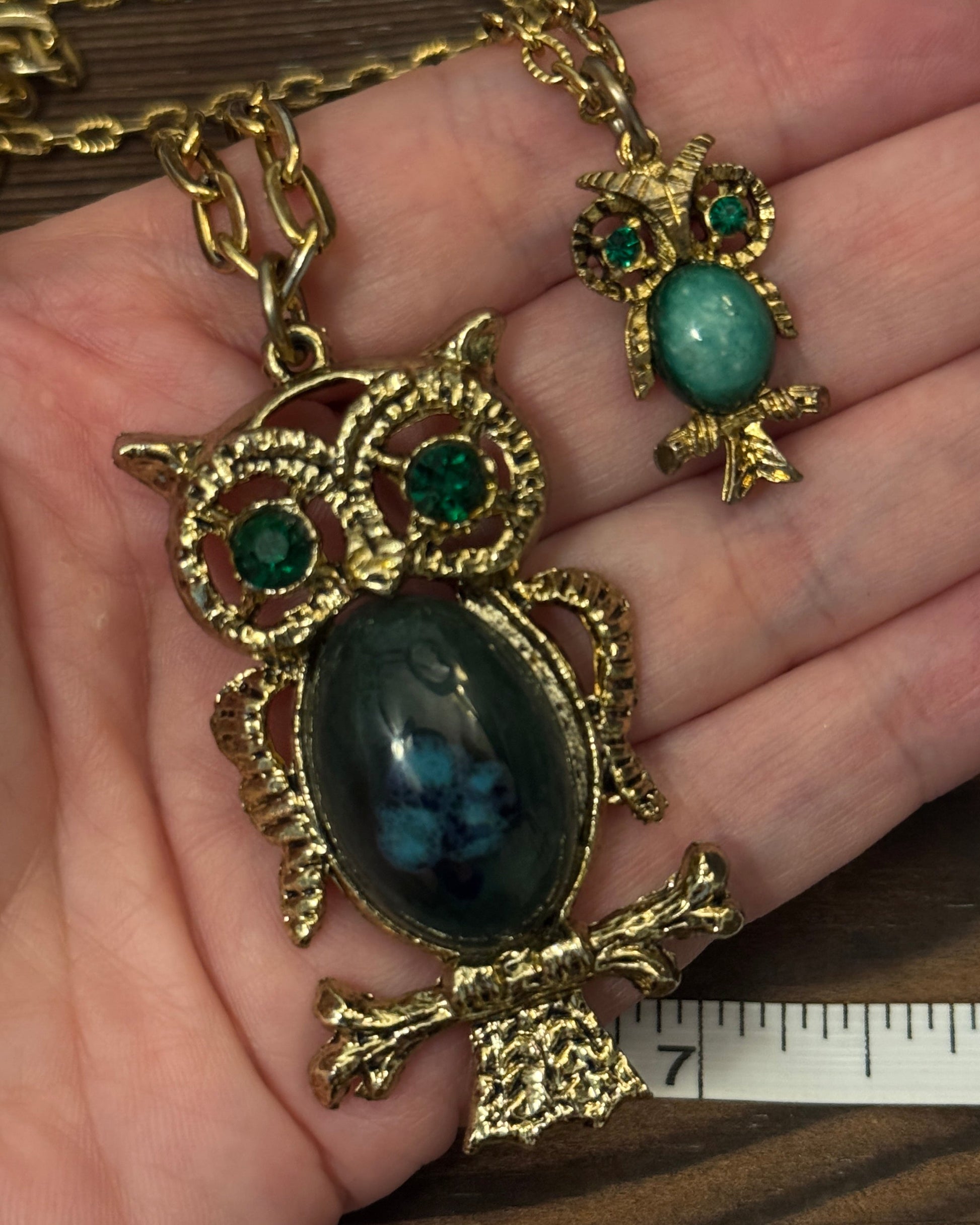 Gold owl-shaped pendant with gemstones held in a hand, next to a ruler for scale.